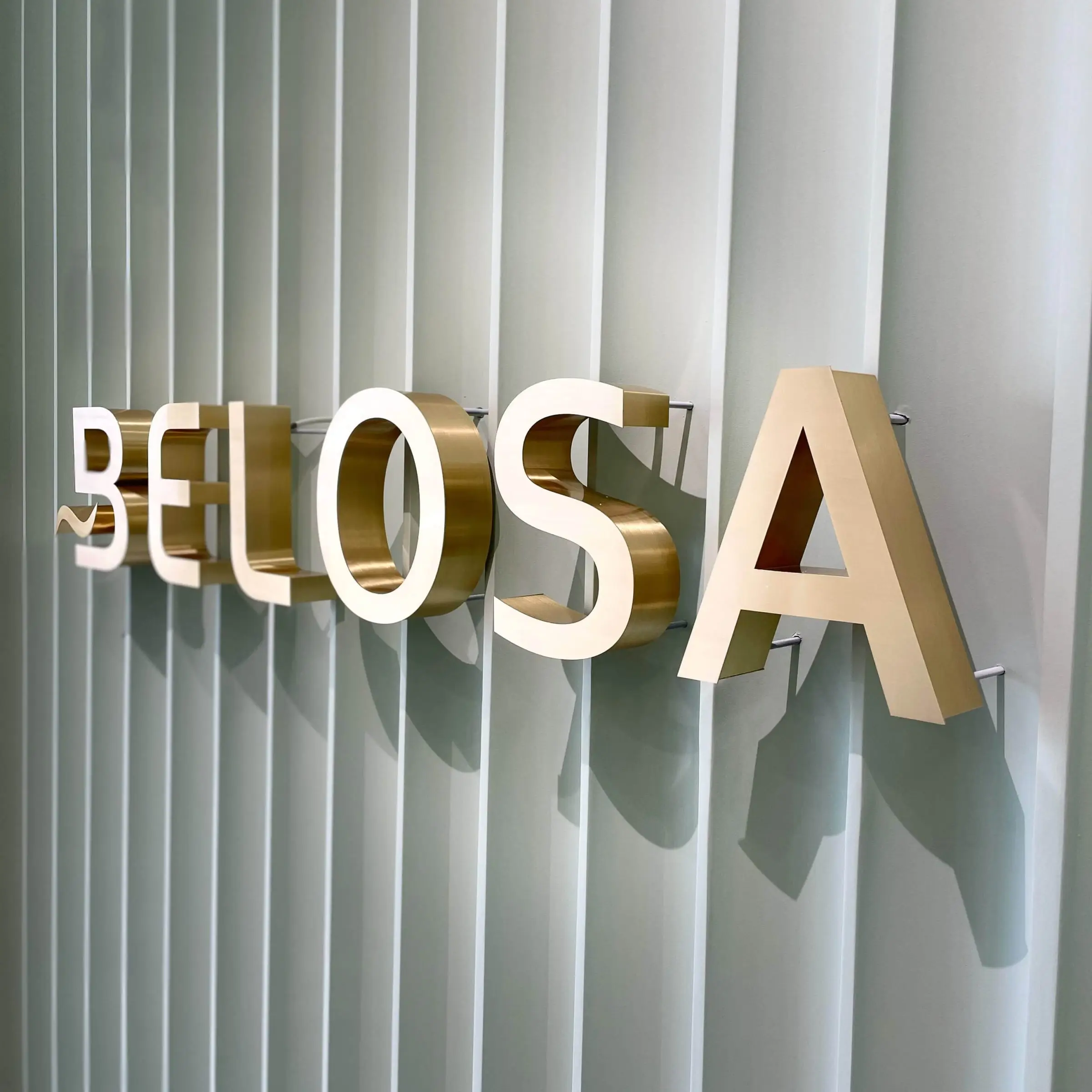 Belosa šveisto žalvario logotipas ant baltos ritmingos sienos