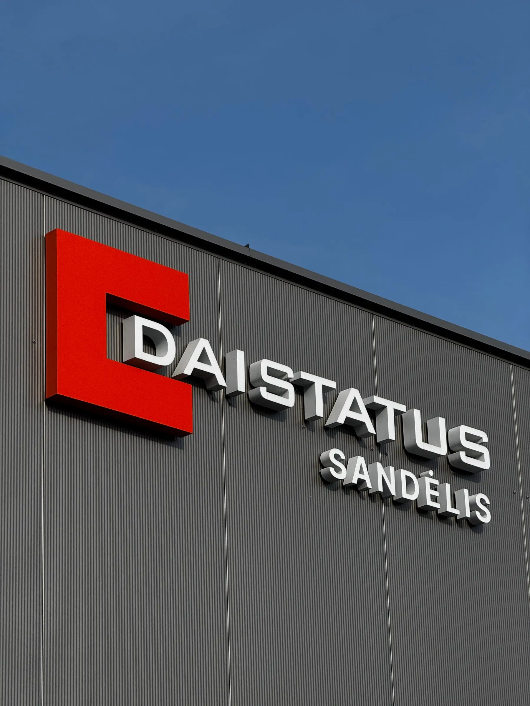 Oranžinis DAISTATUS logotipas ir baltos tūrinės raidės „DAISTATUS sandėlis“ ant pilko sandėlio fasado