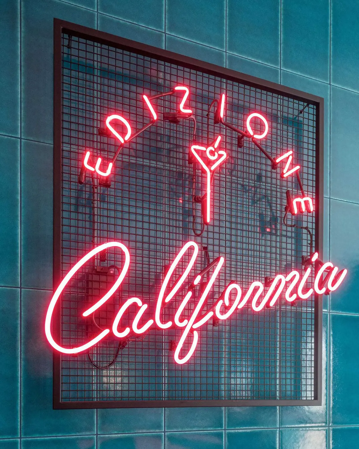 Rožinis neono užrašas „Edizione California“ ant tinklo rėmo ir žalių plytelių